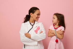 sorridente donna medico e carino bambino ragazza guardare a ogni Altro, indossare rosa nastro, simbolo di Seno cancro consapevolezza giorno, in piedi con attraversato braccia su il petto su colorato sfondo con copia spazio foto