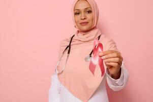 messa a fuoco su Seno cancro consapevolezza nastro, nel mani di sfocato musulmano donna medico nel rosa hijab, isolato su rosa sfondo, copia spazio. 1 st ottobre, mondo giorno di combattimento Seno cancro foto