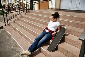 attraente africano americano donna nel casuale capi di abbigliamento in posa su le scale con skateboard e godendo soleggiato giorno foto