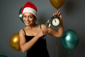 attraente sorridente donna nel Santa cappello celebra Natale e nuovo anno, tintinna con un' Champagne flauto e allarme orologio nel sua mani, guardare carino a il telecamera, in posa su grigio sfondo. copia spazio foto