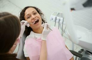 attraente giovane donna con bellissimo Sorridi si siede nel il dal dentista sedia con sua bocca Aperto mentre il igienista esamina o ossequi sua denti utilizzando inossidabile acciaio dentale strumenti - specchio e sonda foto