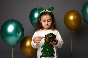 carino bambino ragazza nel elfo cerchio detiene sua regalo nel verde luccichio involucro carta e d'oro nastro, sta contro grigio sfondo con bellissimo brillante verde metallico e d'oro aria palle, copia spazio foto