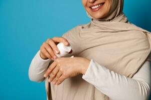 avvicinamento di donna nel hijab spremitura crema su mano, isolato su blu sfondo con spazio per testo foto