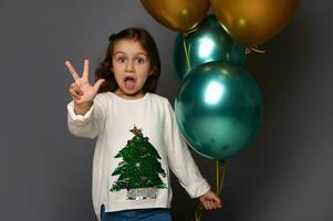 bellissimo ragazza indossare bianca maglione con Natale albero, detiene bellissimo d'oro e verde metallico aria palloncini nel sua mano, Spettacoli dita guardare a telecamera, pose contro grigio sfondo, copia spazio foto