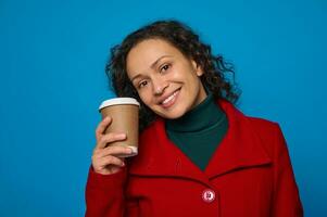 affascinante dai capelli scuri bella donna nel verde Maglione e luminosa rosso cappotto detiene un' cartone monouso tazza con caldo bevanda e sorrisi allegramente in posa contro blu sfondo con copia anno Domini spazio foto