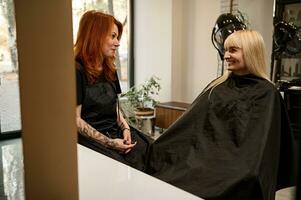 alla moda bella dai capelli rossi donna nel nero professionale uniforme, capelli stilista con elegante corpo tatuaggi su braccia parlando con un' capelli salone cliente seduta nel barbiere sedia nel davanti di un' specchio foto