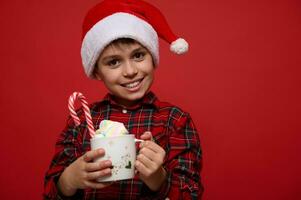 adorabile bambino ragazzo con bellissimo trentadue denti Sorridi, indossare Santa cappello e in posa contro rosso sfondo con un' tazza di caldo cioccolato bevanda con marshmallows e a strisce dolce caramella canna. Natale concetto foto