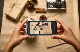 umano mani Tenere mobile Telefono nel vivere Visualizza modalità e assunzione un' foto di Natale composizione di tazza con cacao e marshmallows, Vintage ▾ telecamera, mestiere regalo scatola e pino coni su cartone superficie