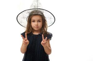 isolato vita lunghezza ritratto di bella poco strega ragazza indossare un' procedura guidata cappello e vestito nel elegante carnevale vestire, gesticolando, mostrando un' pace cartello con dita. Halloween concetto foto