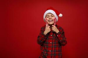 bellissimo tenero carino poco ragazzo, adorabile sorridente bambino nel Santa cappello e scacchi camicia delicatamente abbracci Natale Lecca-lecca dolce a strisce caramella canne, isolato su rosso sfondo, copia spazio per anno Domini foto