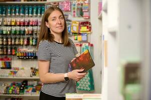 piacevole femmina venditore detiene un' taccuino, sorrisi guardare a telecamera mentre lavori su merchandising nel scuola Stazionario negozio foto