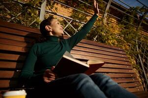 rilassato contento giovane donna nel casuale denim saluto con mano, lettura un' libro seduta su un' di legno panchina godendo un' caldo soleggiato autunno fine settimana nel nazione parco, lontano a partire dal urbano spingere foto