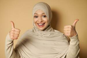 contento arabo musulmano donna nel hijab con un' bellissimo trentadue denti Sorridi mostrando pollici su per telecamera. beige sfondo con copia spazio. foto