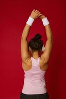ritratto di femmina atleta nel abbigliamento sportivo e senza fili cuffie fare allungamento esercizi su braccia in piedi indietro per il telecamera contro rosso sfondo foto