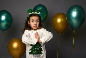 bellissimo poco ragazza nel Natale maglione e elfo cerchio pose con mani palma per palma, sembra a telecamera, isolato al di sopra di grigio sfondo con d'oro metallico verde aria palloncini con copia spazio per anno Domini foto