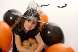 ritratto di un' bellissimo poco ragazza indossare un' procedura guidata cappello, vestito nel buio strega carnevale costume, abbracci un' zucca nel sua mano, sedersi su un' bianca sfondo con colorato nero arancia palloncini e pipistrelli foto