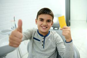 adorabile bello bambino ragazzo sorrisi con bellissimo trentadue denti Sorridi e Spettacoli pollice su e giallo visitare carta con copia anno Domini spazio, guardare a telecamera mentre seduta nel dal dentista sedia dopo dentale trattamento foto