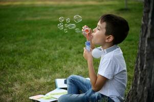 adorabile scuola ragazzo godendo il suo ricreazione fra classi, soffiaggio sapone bolle, seduta su il verde erba di urbano città parco Il prossimo per il suo dire bugie giù scuola forniture e Accessori foto