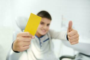 messa a fuoco su vuoto giallo visitare carta con copia spazio per inserire Annunci e testo nel il mano di sfocato bello ragazzo sorridente trentadue denti Sorridi e mostrando pollice su guardare a telecamera seduta nel dentista sedia foto