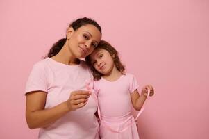 adorabile bambino ragazza , Leans su sua madre spalla, entrambi vestito nel rosa colore vestito, hold rosa raso nastri, carino Sorridi guardare a telecamera . mondo Seno cancro consapevolezza giorno. combattimento cancro. copia spazio foto