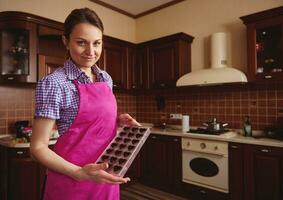 cioccolatiere in posa su telecamera con muffe pieno di cioccolato conchiglie su il sfondo di di legno casa cucina foto