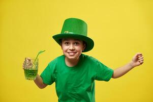 contento ragazzo nel fata cappello con mani su detiene un' bicchiere con verde bevanda e esprimere felicità festeggiare il santo Patrick giorno. copia spazio. giallo sfondo foto