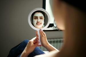 riflessione su il specchio di un' sorridente donna con alginato maschera su sua viso guardare a se stessa nel un' poco specchio. pelle cura concetto foto