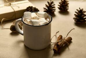 legato cannella nel arco a partire dal un' corda Il prossimo per semplice rustico vecchio tazza di caldo cacao bevanda e bianca dolce marshmallows su un' cartone sfondo con pino coni e Natale regalo nel mestiere involucro carta foto