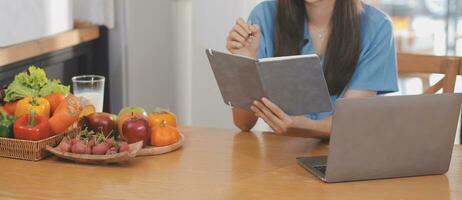 un' giovane donna con un' bellissimo viso nel un' blu camicia con lungo capelli mangiare frutta seduta dentro il cucina a casa con un' il computer portatile e taccuino per rilassamento, concetto vacanza. foto