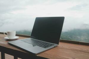 computer tenere sotto controllo, tastiera del computer, caffè tazza e topo con vuoto è su il opera tavolo a il cielo montagna fiume e alberi davanti Visualizza sfondo. foto