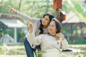 asiatico anziano donna nel sedia a rotelle con contento figlia. famiglia relazione pensionato donna seduta su sedia a rotelle nel il parco età cura a la pensione casa. foto