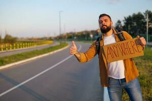 uomo è autostop su ciglio della strada provando per fermare macchina. lui è Tenere cartone con iscrizione. foto