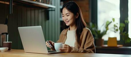 asiatico donna seduta a scrivania nel cucina godendo caffè spuntini e in linea soddisfare foto