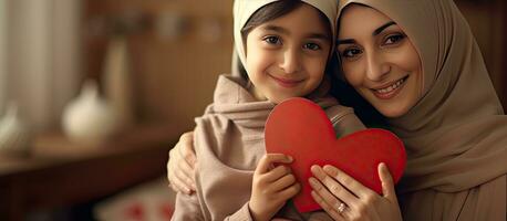 poco ragazza dando madre un' regalo cuore disegnato su saluto carta madre indossare hijab contento bambino abbracciare madre e sorridente a il telecamera foto