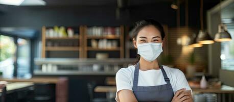 asiatico femmina barista nel piccolo attività commerciale istituzione indossare viso maschera durante covid 19 pandemia in posa con fiducia con braccia attraversato e un' allegro smi foto