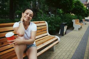 attraente femmina turista sorridente a telecamera, Tenere carta tazza con porta via caffè, seduta su di legno panchina nel il parco foto