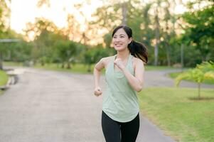 in forma asiatico giovane donna jogging nel parco sorridente contento in esecuzione e godendo un' salutare all'aperto stile di vita. femmina pareggiatore. fitness corridore ragazza nel pubblico parco. salutare stile di vita e benessere essere concetto foto