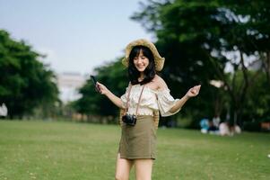 ritratto di asiatico giovane donna viaggiatore con tessitura cappello, cestino, mobile Telefono e telecamera su verde pubblico parco sfondo. viaggio viaggio stile di vita, mondo viaggio esploratore o Asia estate turismo concetto. foto