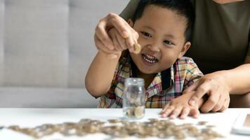 contento asiatico ragazzo e mamma Salvataggio i soldi insieme, mettendo denaro contante in porcellino banca. madre giocando con bambino su riscaldamento pavimento a casa, insegnamento poco figlio per investire i soldi, pianificazione futuro. foto