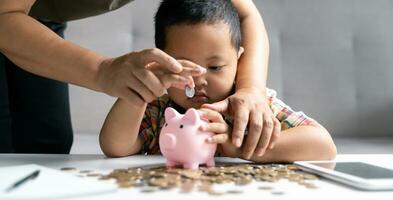 contento asiatico ragazzo e mamma Salvataggio i soldi insieme, mettendo denaro contante in porcellino banca. madre giocando con bambino su riscaldamento pavimento a casa, insegnamento poco figlio per investire i soldi, pianificazione futuro. foto