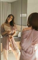 contento mattina. attraente giovane donna guardare nel specchio a sua appartamento - scia su e nuovo giorno foto