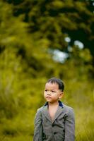 carino poco ragazzo un' ben vestito ragazzo nel un' completo da uomo nel un' Giardino dietro la casa con un' prato e guardare per qualcosa interessante. foto