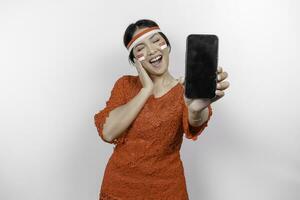 un' ritratto di un' sorridente asiatico donna indossare rosso kebaya e fascia per capelli e mostrando sua Telefono, isolato di bianca sfondo. dell'Indonesia indipendenza giorno concetto foto