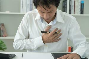 mano hold il petto con cuore attacco sintomi, asiatico uomo Lavorando difficile avere il petto dolore causato di cuore patologia, perdere, dilatazione, allargata coronario cuore, stampa su il il petto con un' doloroso espressione foto