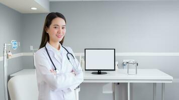professionale giovane asiatico donna medico indossare bianca accappatoio e stetoscopio in piedi con braccia attraversato contento e sorridi a visita medica camera nel Ospedale. foto