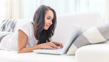 attraente sorridente giovane femmina nel un' bianca maglietta e pantaloni della tuta dire bugie su un' divano, scrittura su un' il computer portatile Appoggiato contro il cuscino foto