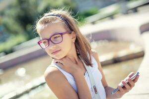 giovane pre-teen bionda ragazza detiene mobile Telefono nel sua mano seduta su il bordo di un' Fontana su un' soleggiato estate giorno foto