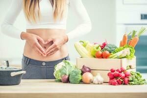 donna con gli sport figura su sua pancia Spettacoli cuore forma nel casa cucina con di legno scatola pieno di biologico verdura foto