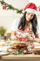 intelligente brunetta nel un' Natale grembiule e Santa cappello prepara un' arrosto Oca o tacchino, strofina esso con miele prima il finale cottura al forno foto