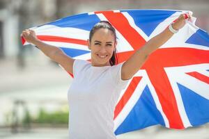 bella ragazza in un bianca maglietta detiene un' bandiera di grande Gran Bretagna dietro a suo, sorridente a il telecamera foto